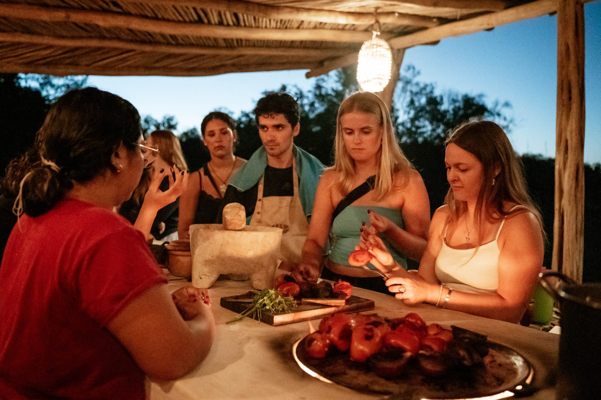 Grupo pequeño preparando chiles y jitomates asados en la mesa de piedra al anochecer